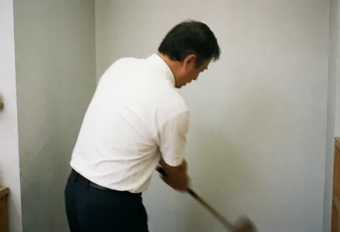 Instructeur de golf en mouvement réalisant un swing complet avec un club de type bois sur fond gris clair, vêtu de blanc et noir.