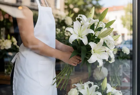 Une fleuriste arrange un bouquet de lys blancs et de feuillage vert, visible à travers la vitrine du magasin depuis l'extérieur.
