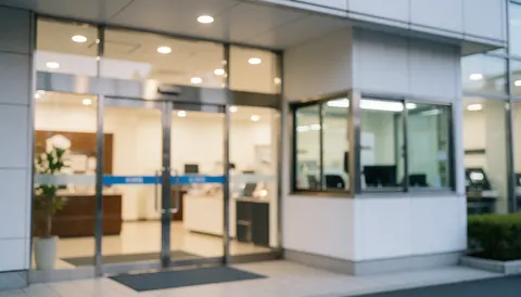 Entrée d'un bâtiment moderne avec des portes vitrées, un agent de sécurité présent et une lumière intérieure chaleureuse.