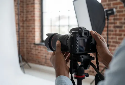 Gros plan des mains d'un photographe réglant la mise au point d'un boîtier DSLR fixé sur un trépode dans un studio avec mur en briques et softbox.