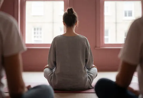 Vue arrière d'une instructrice de yoga assise en position de lotos dans un studio aux murs roses et à la lumière naturelle.
