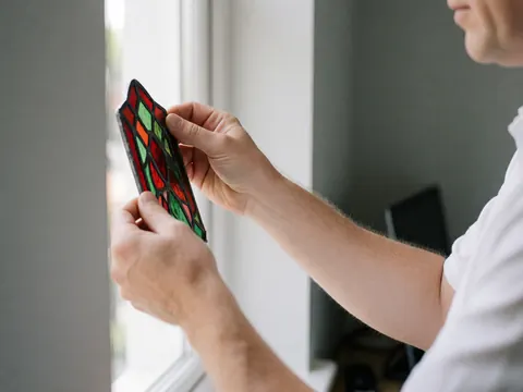 Des mains d'artisan vitrier ajustent un fragment de verre aux motifs rouges et verts contre la lumière naturelle dans un atelier aux murs gris.