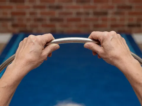 Des mains adultes saisissent un anneau métallique au-dessus d'une grille bleue, avec un mur de briques rouges en arrière-plan, évoquant un entraîneur de gymnastique.
