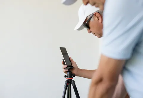 Instructeur de golf professionnel tenant un téléphone portable fixé sur un trépied devant un mur blanc, éclairage naturel et réaliste.