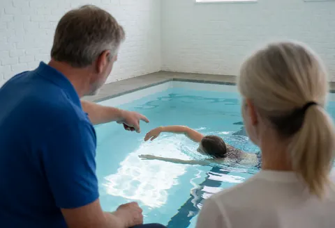 Un entraîneur de natation donne des instructions à un étudiant dans une piscine intérieure aux murs en briques blanches, avec un geste d'enseignement visible.