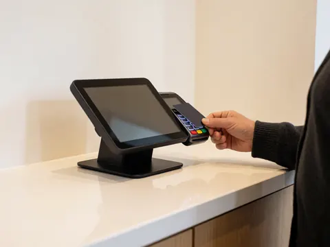 Une main effectue un paiement par carte sur une borne de paiement posée sur un comptoir propre, devant des murs blancs et sous une lumière vive.