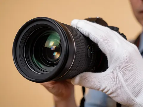 Une main portant un gant blanc ajuste avec précision la bague de mise au point sur un objectif de caméra professionnel, posé sur une surface beige.