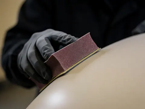 Main portant un gant noir utilisant un bloc de ponçage à grain 800 sur un panneau courbé de voiture dans un atelier sombre avec une lumière focalisée.