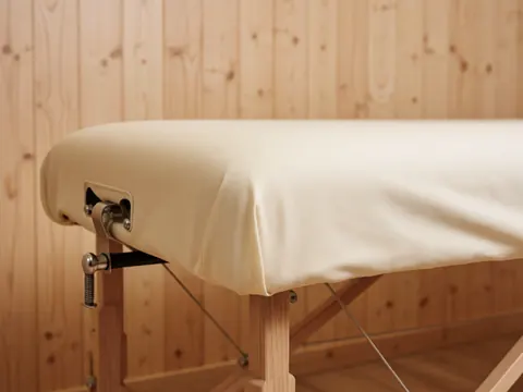 Vue rapprochée des attaches métalliques fixées au coin d'une table de massage recouverte d'un tissu en coton, avec un mur en bois naturel et une lumière douce.