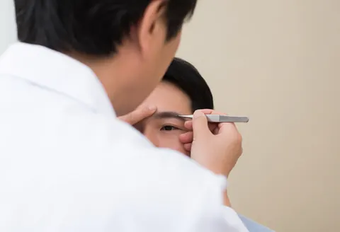 Un spécialiste réalise un microblading sur la sourcil d'un client à l'aide de pinces, sur fond beige clair et sous une lumière vive.
