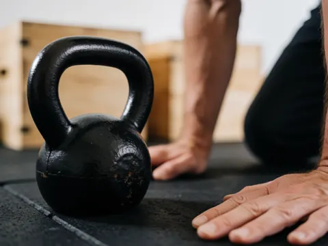 Kettlebell noir en fonte texturée posé sur un tapis de sol en caoutchouc, mains d'adulte au premier plan, arrière-plan en bois, idéal pour un coach de CrossFit.