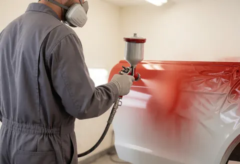 Un technicien en tenue de protection peint une portière de voiture avec une bombe de peinture rouge dans un atelier aux murs blancs.