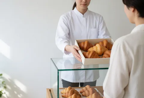 Un boulanger en uniforme blanc sert une boîte de croissants dans une vitrine en verre, avec une lumière naturelle et un mur blanc en arrière-plan.