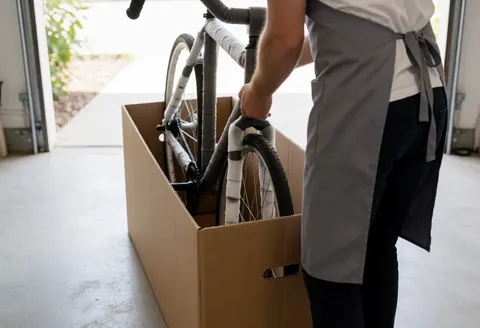 Un professionnel portant un tablier gris empaquete un vélo démonté dans une caisse en carton, au sein d'un garage éclairé par la lumière du coucher de soleil.