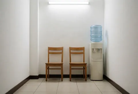 Un coin repos avec deux chaises en bois et un distributeur d'eau dans un dojo aux murs blancs et au sol carrelé.
