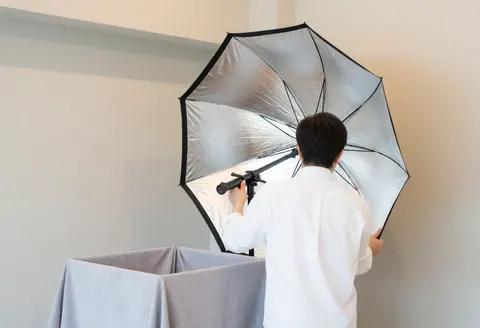 Un photographe ajuste un réflecteur en forme de parapluie près d'une boîte bébé avec tissu gris dans un studio aux murs neutres et à l'éclairage doux.