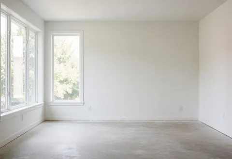 Espace intérieur vide aux murs blancs et au sol en béton, éclairé par la lumière naturelle entrant par de grandes fenêtres, idéal pour l'installation de revêtements.