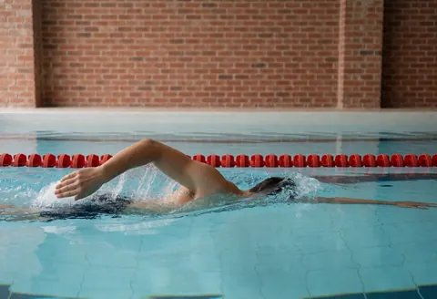 Un nageur professionnel effectuant un crawl dans une piscine intérieure avec des murs en briques et une ligne rouge visible.