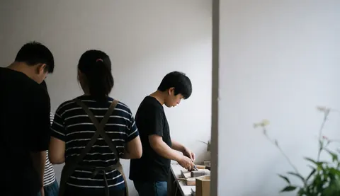 Trois individus regardent un artisan utiliser des outils dans un atelier de menuiserie aux murs blancs.
