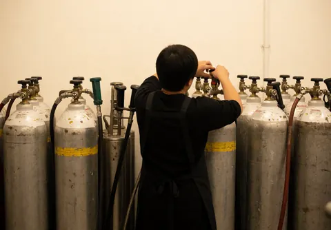 Personne en tablier noir ajustant une vanne sur un réservoir de gaz parmi d'autres, dans un local aux murs blancs, pour un décorateur d'événements.