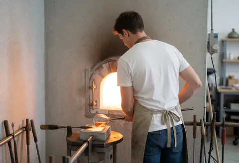 Un artisan travaille le verre en fusion à l'aide d'outils et de moules dans un atelier industriel.