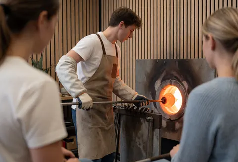 Un artisan souffleur de verre manipule un tube contenant du verre en fusion devant une gloria, dans un atelier aux murs en bois clair.