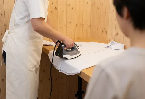Un tailleur portant un tablier blanc repasse une couture de chemise blanche sur une planche en bois avec des murs en panneaux naturels.