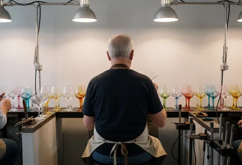 Un artisan souffleur de verre travaille dans son atelier avec des verres colorés posés sur des étagères, sous l'éclairage de travail, devant des murs gris.