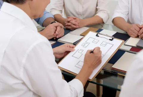 Un professionnel dessine des plans de mobilier sur une table en verre avec des échantillons de tissu pendant une réunion client.