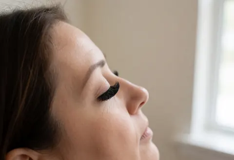 Fermeture des yeux mettant en valeur de longs cils postiches dramatiques, éclairée par une lumière naturelle douce devant un mur beige flou dans un salon de beauté.
