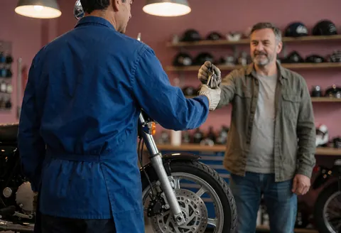 Un mécanicien professionnel remet les clés à un client satisfait dans un atelier de réparation de motos, avec une roue avant visible et des casques sur des étagères en arrière-plan.