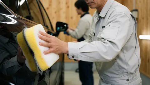 Un employé en uniforme nettoie un véhicule sombre à l'aide d'une éponge jaune dans un atelier aux murs en bois clair.