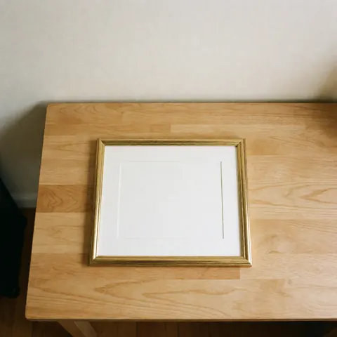 Cadre doré pour diplôme posé sur une table en bois clair, fond de mur blanc chaud, présentation propre et minimaliste pour photographe de cérémonies.