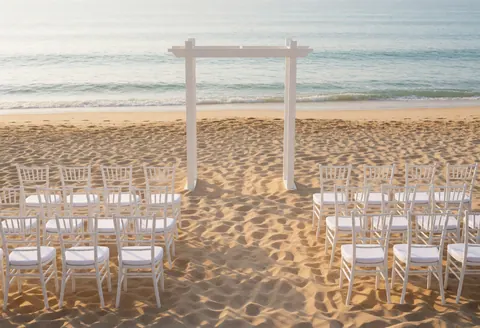 Un arc blanc décoratif posé sur le sable face à l'océan, entouré de chaises blanches pour une cérémonie, baigné par une douce lumière du matin.