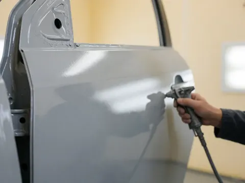 Une main tient un pistolet de pulvérisation pour appliquer une base grise sur la portière d'un véhicule dans un atelier aux murs clairs.