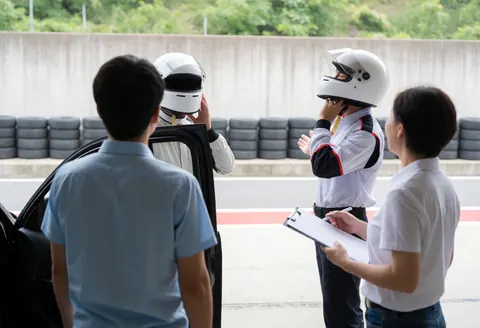 Un marshal avec un bloc-notes examine un pilote en tenue de course près des barrières de pneus et de béton sur un circuit de sport automobile.