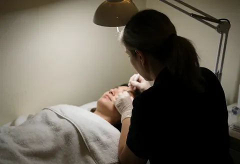 Une esthéticienne en gants applique un sérum hydratant sur la zone oculaire d'une cliente allongée sur une serviette blanche dans un salon aux tons chauds.