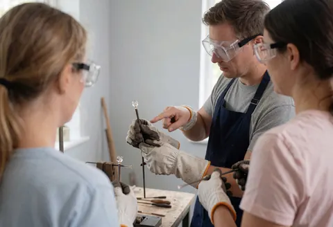 Instructeur de verrerie avec des lunettes et des gants enseignant à des étudiants à modeler du verre chaud avec des outils dans un atelier bien éclairé.