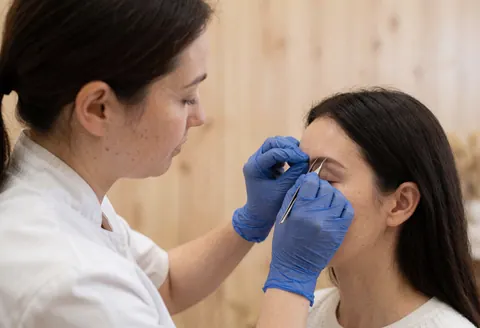 Une technicienne portant des gants bleus applique des faux cils à un client aux yeux fermés dans un salon de beauté au mur en bois clair.
