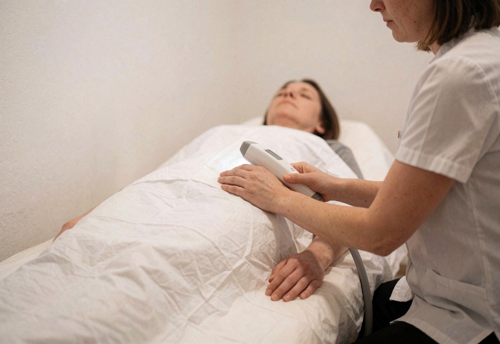 Une praticienne en uniforme effectue une épilation au laser sur un client posé sur une table de soin dans un salon clair et calme.
