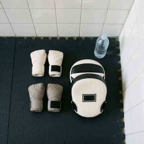 Gants de boxe accompagnés de bandages et de masques d'entraînement posés sur une natte noire devant un mur de carreaux blancs.