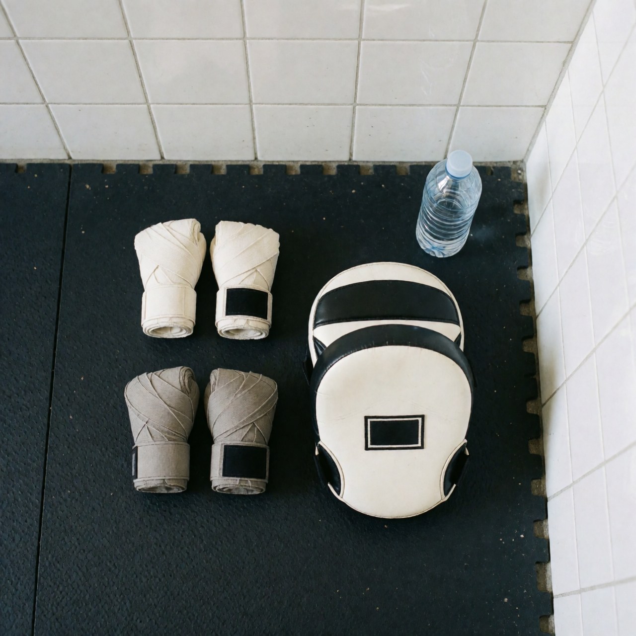Gants de boxe accompagnés de bandages et de masques d'entraînement posés sur une natte noire devant un mur de carreaux blancs.