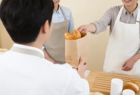 Un employé en tablier tend un emballage de pain frais à un client heureux dans une boulangerie éclairée par la lumière naturelle.
