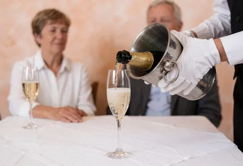 Un serveur verse du champagne dans des flûtes posées sur une table blanche avec des nappes, tandis que des clients sont assis en arrière-plan lors d'une célébration.