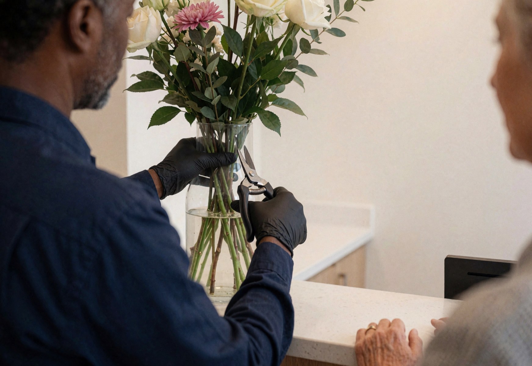 Une floriste aux gants coupe des tiges de roses blanches et roses dans un vase en verre sur le comptoir, avec une cliente à côté.