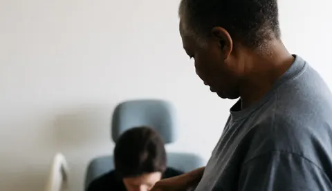 Un thérapeute effectue un massage à un adulte assis sur une chaise pliante dans un intérieur lumineux avec un mur blanc.