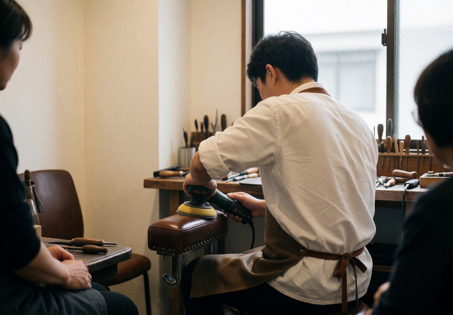 Un tapissier portant un tablier en cuir utilise un outil électrique pour poncer un siège dans un atelier aux murs blancs et étagères.