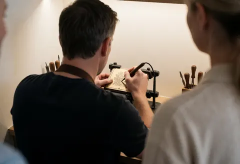 Un artisan sculpte un motif sur une planche en bois dans son atelier, entouré d'outils et de murs blancs.