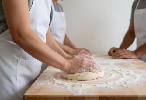 Des mains en train de pétrir de la pâte sur une planche à pain en bois, parsemée de farine, dans un cadre lumineux et épuré.