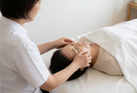 Une praticienne applique des gestes doux de shiatsu sur le visage d'un client allongé sur une table de massage dans un espace lumineux et apaisant.